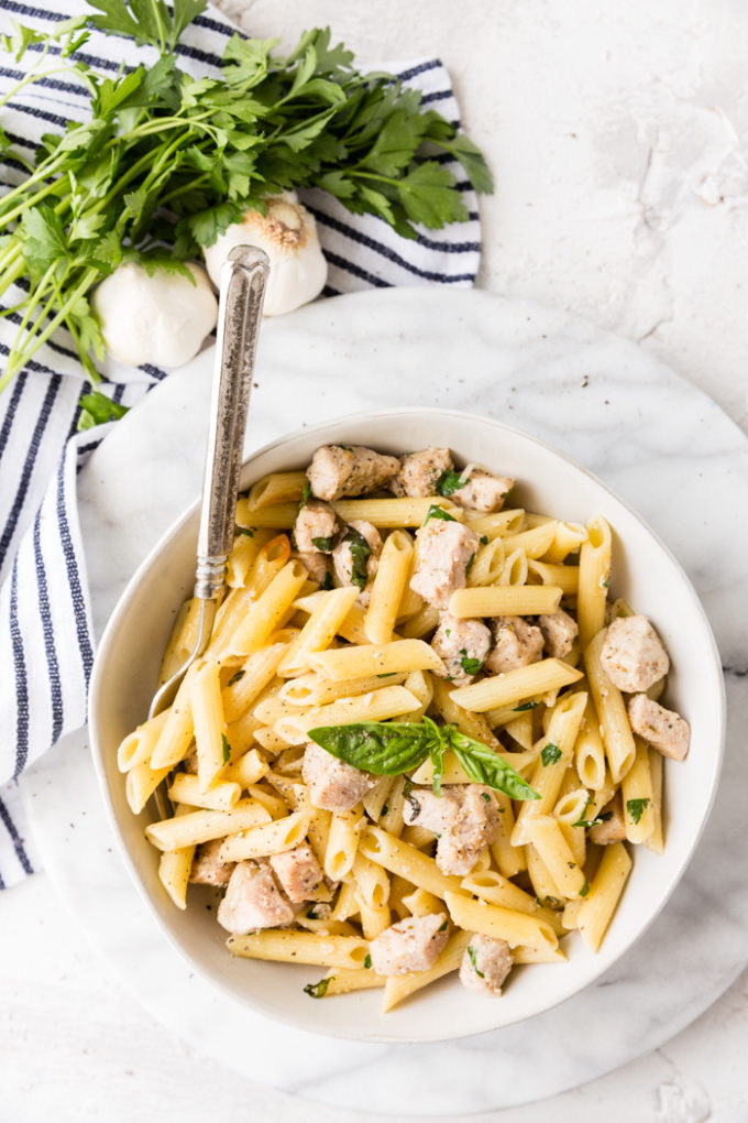 Roasted garlic and herb penne pasta in a white bowl with a blue and white napkin, cloves of garlic, and fresh parsley