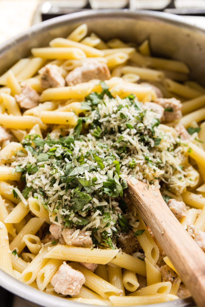 A pot of penne pasta with a pile of herbs and seasonings on top ready to be mixed in with a wooden spoon