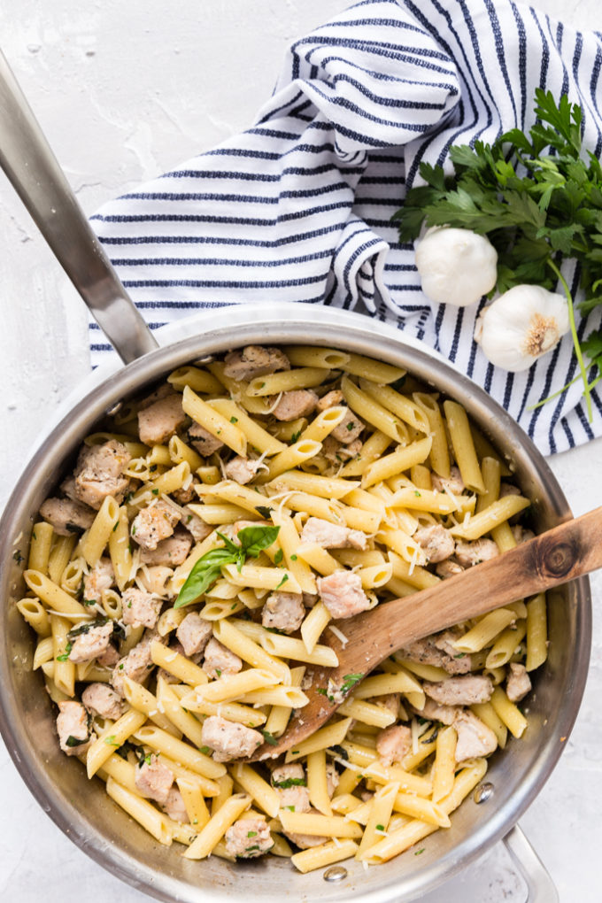 A skillet of roasted garlic and herb penne pasta with pork, on a blue and white striped napkin with garlic cloves and fresh herbs