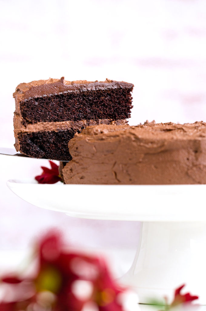 A chocolate cake on a white pedestal, with one slice cut out and half way lifted off the stand. Two layers of chocolate cake with a chocolate buttercream icing. 