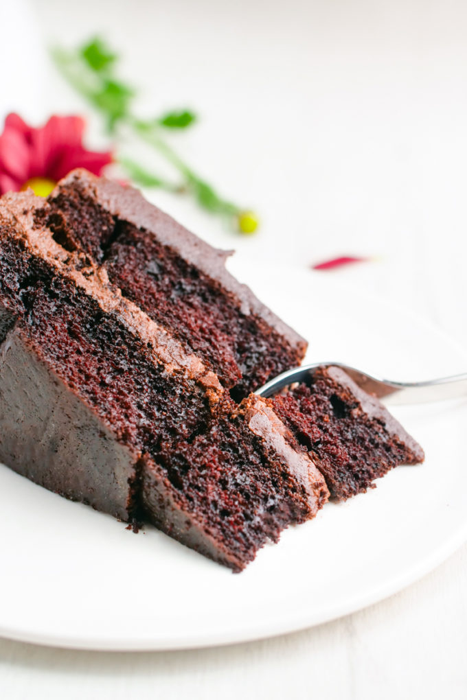 A slice of double layered chocolate cake with chocolate buttercream icing on a white plate. 