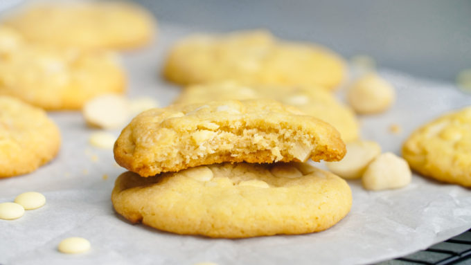A delicious white chocolate macadamia nut cookie with a bite out, sitting a top another white chocolate chip macadamia nut cookie with a few cookies in the background