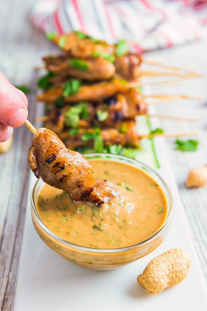 Chicken satay being dipped in a glass bowl of peanut dipping sauce. 