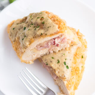 White plate with homemade chicken cordon bleu, cut in half, topped with a creamy sauce and garnish, fork resting on plate