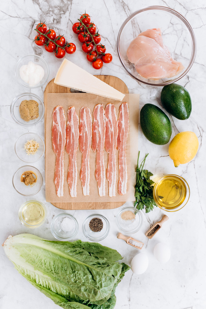 All the ingredients needed for a chicken avocado caesar salad, bacon, lettuce, avocado, lemon, chicken tomatoes, all on a marble surface