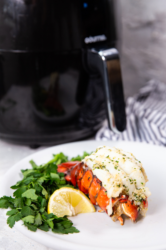 Air fryer lobster tail on a white plate with parsley and lemon slice with an air fryer behind the plate