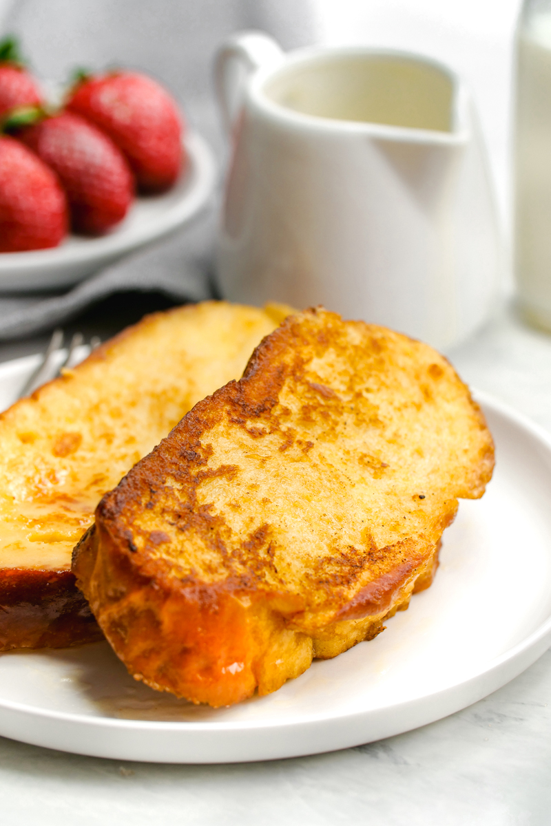 White plate with two slices of french toast, strawberries in the background, and a jug of syrup