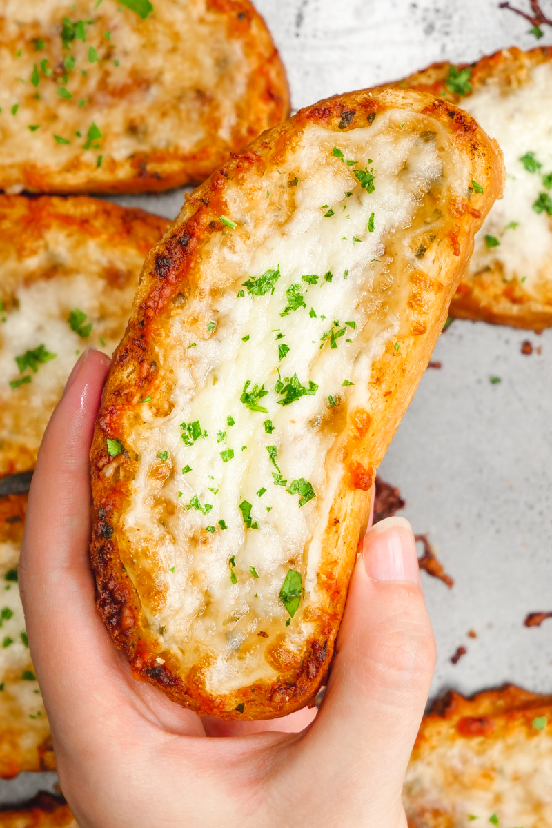 A lovely slice of cheesy garlic bread in a hand over a sheet of garlic bread