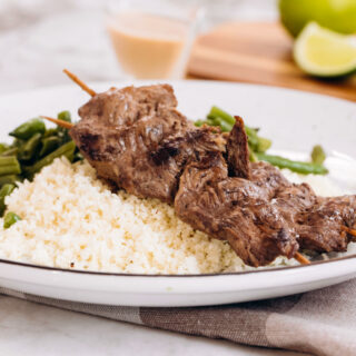 A white plate sitting on top of a checked napkin, topped with cauliflower rice, beef satay, and green beans