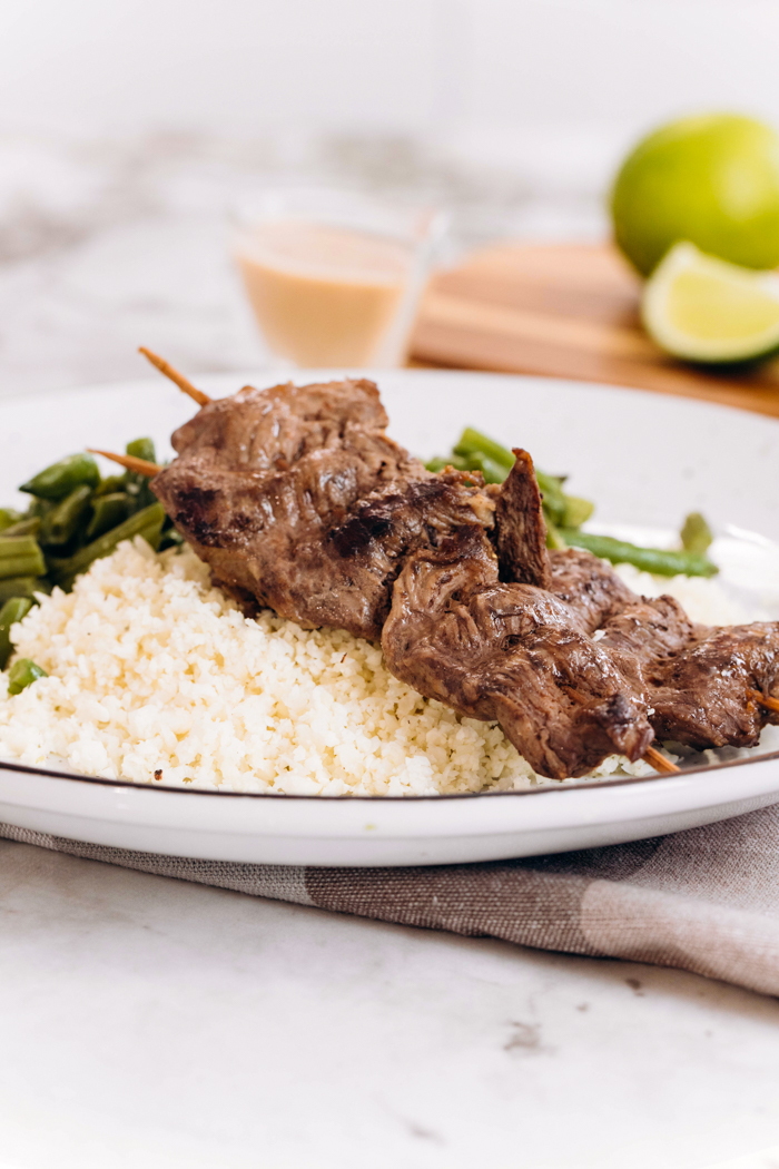 A white plate sitting on top of a checked napkin, topped with cauliflower rice, beef satay, and green beans