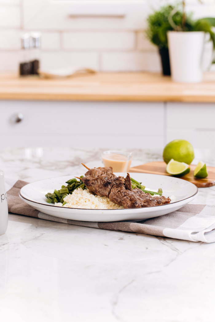 Low carb beef satay on a white plate with a checked napkin, some peanut sauce, limes. and a shelf in the background