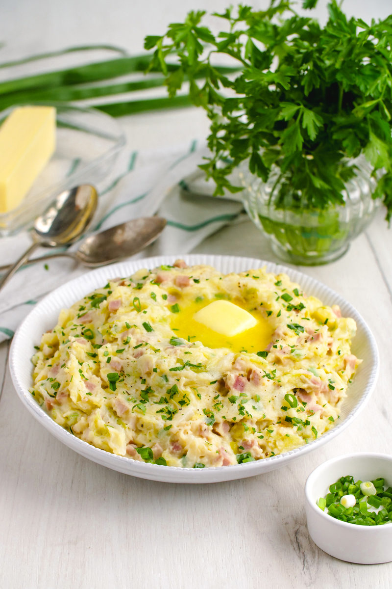 A white bowl filled with colcannon and topped with a pad of butter. Two spoons behind the colcannon and a vase of fresh parsley