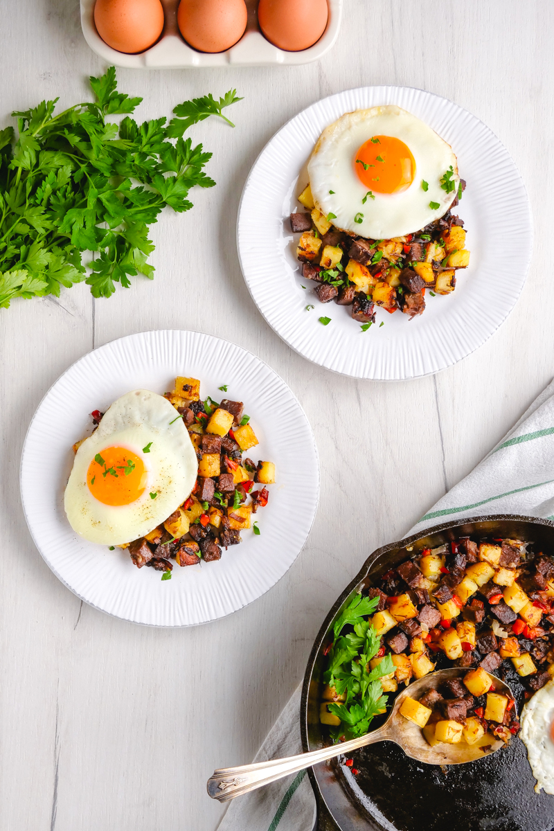 Two white plates topped with corned beef hash and eggs, with fresh parsley and eggs in the background