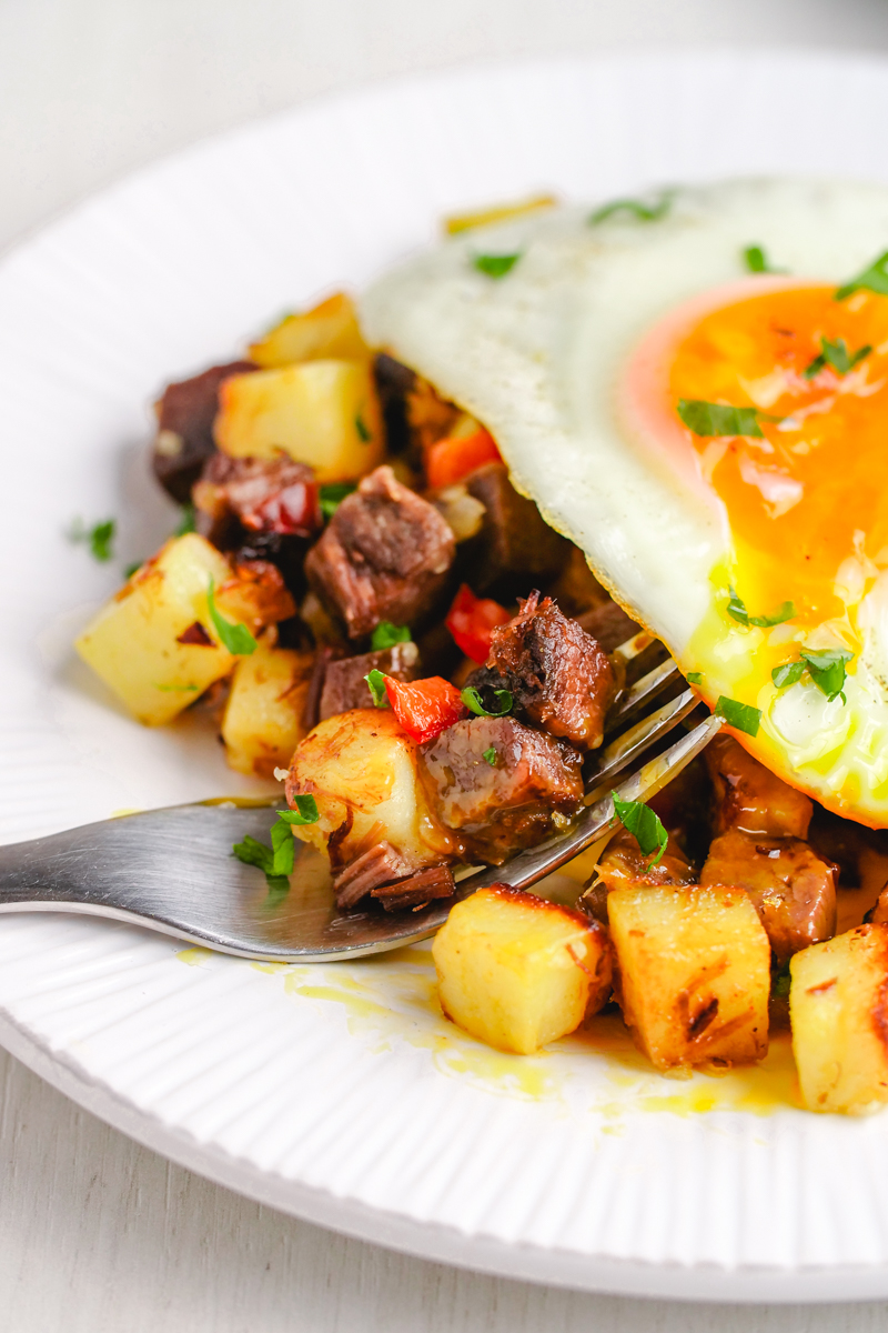 A close up of corned beef hash with an egg on top on a white plate with a fork holding some of the corned beef hash recipe