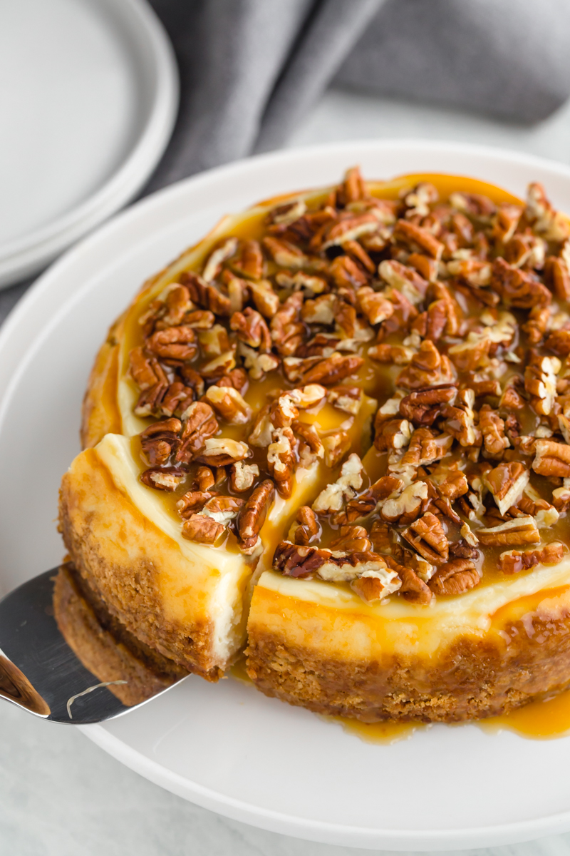 A whole instant pot cheesecake topped with caramel and pecans, with a slice cut and partially pulled out. 