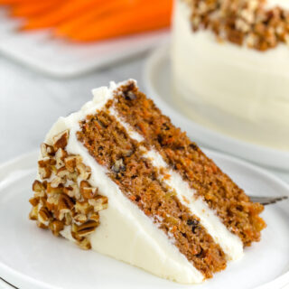 Carrot cake slice with cream cheese icing on a white plate with carrots in the background