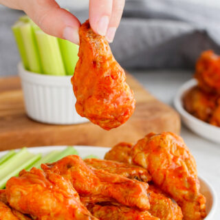 Buffalo wings, these hot wings are on a white plate with celery in the background, and a hand holding one chicken wing up.