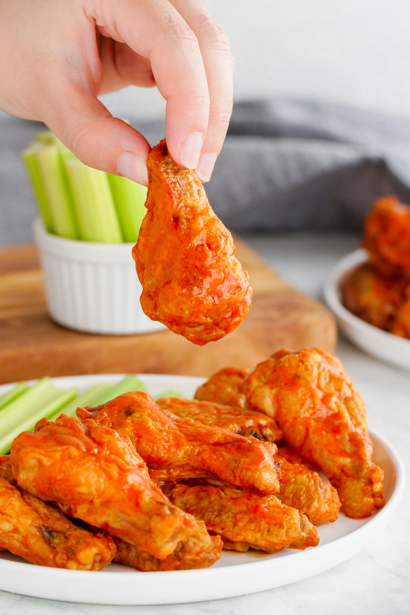 Buffalo wings, these hot wings are on a white plate with celery in the background, and a hand holding one chicken wing up.