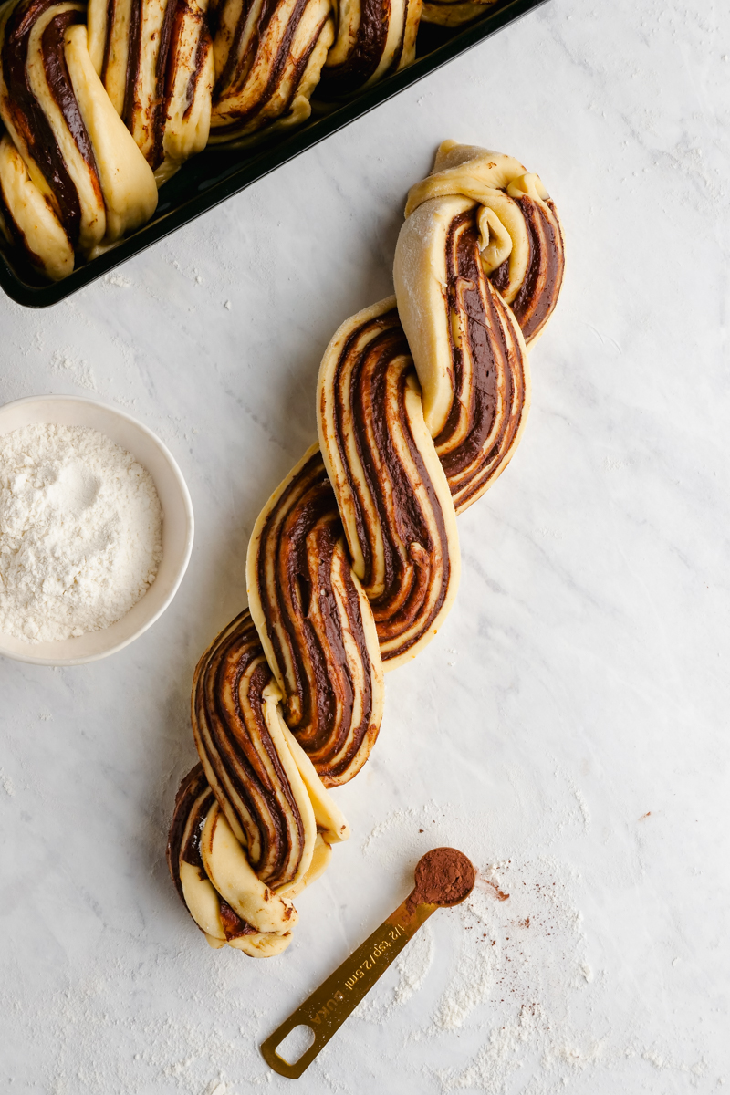 A braid of babka, with a measuring spoon and a portion bowl, and a pan holding babka above it. 