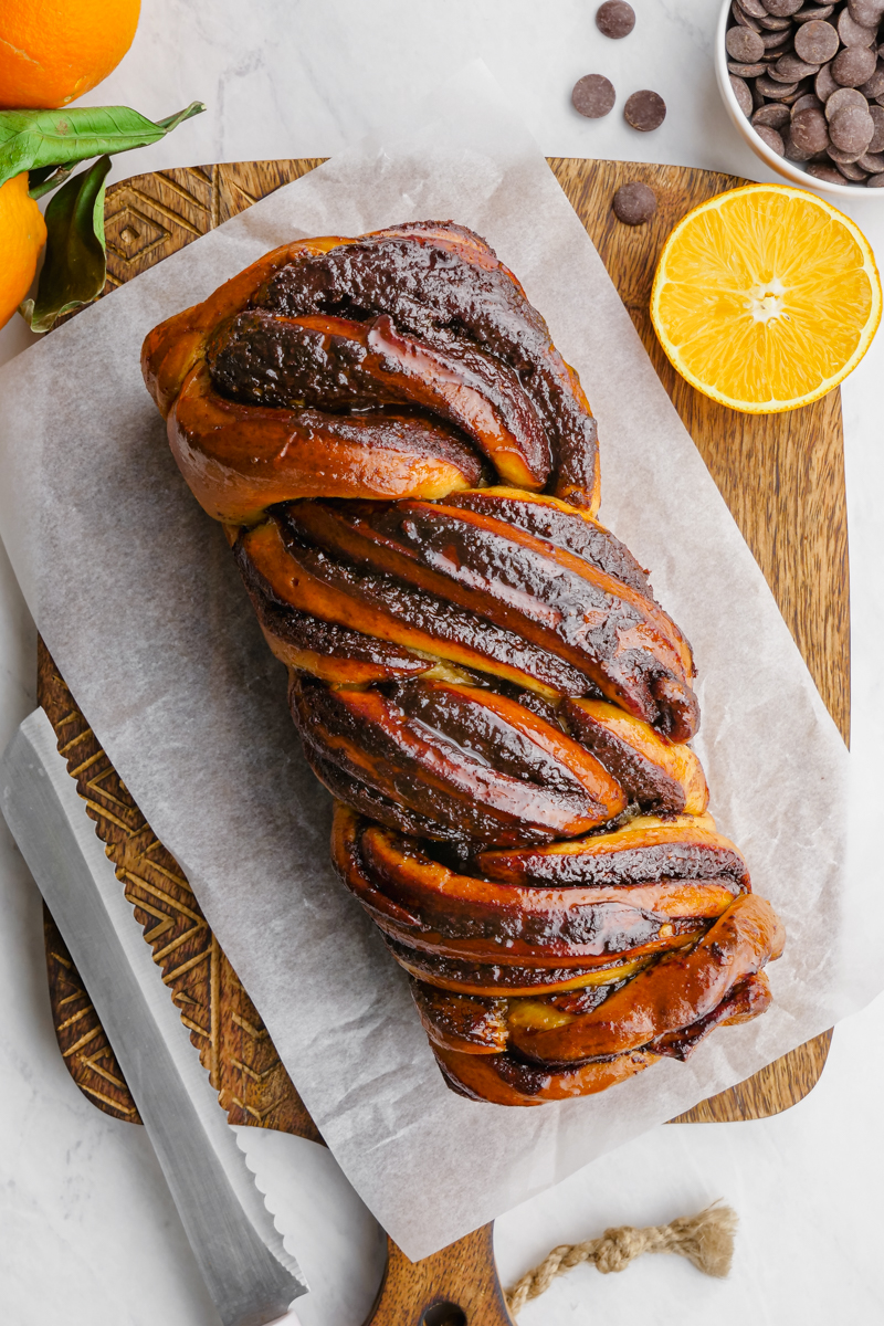 A top down look at a loaf of babka on top of a piece of parchment on a wooden cutting board. Knife next to the loaf. ORanges and chocolate garnish
