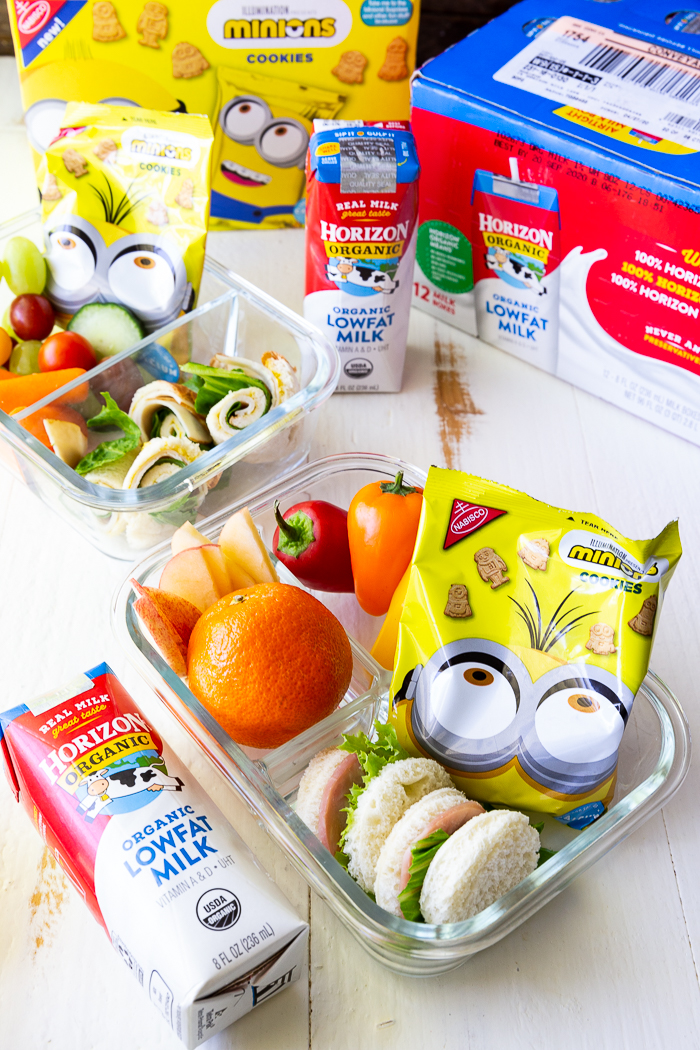 glass bento box with mandarin orange, grape tomatoes, carrot sticks and celery sticks, a round sandwich and a yellow package of Nabisco minion cookies, packaging for horizon organic low fat milk boxes and Nabisco Minion cookies behind