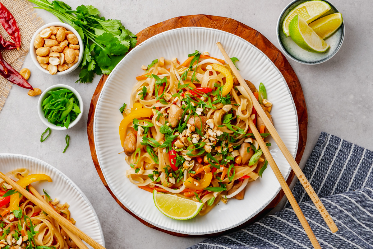 A top down image of a white plate with pad thai, chop sticks, lime wedges, and other garnish