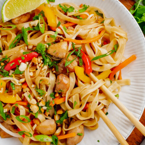 A white plate with pad thai, chopsticks, a lime, and toppings