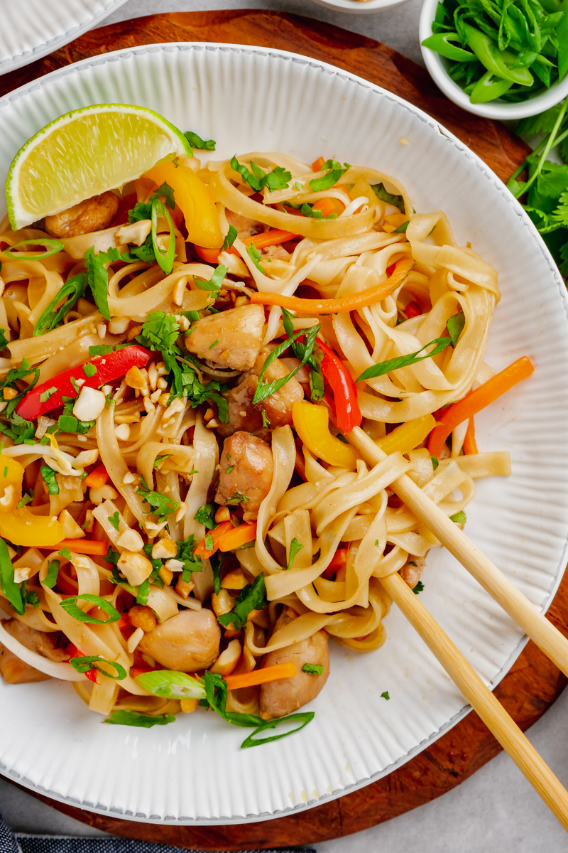 A white plate with pad thai, chopsticks, a lime, and toppings