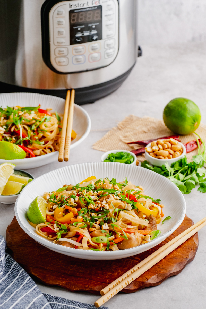 An instant pot in the background, bowls of pad thai and chop sticks, as well as garnish
