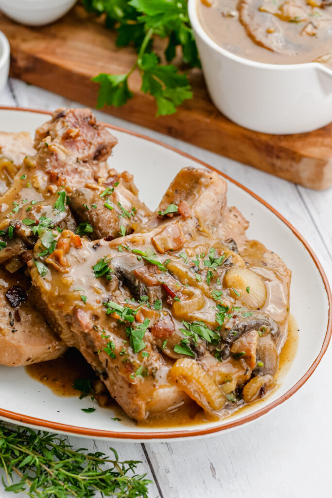 Smothered pork chops on a white serving dish with a cutting board and gravy in the background. 