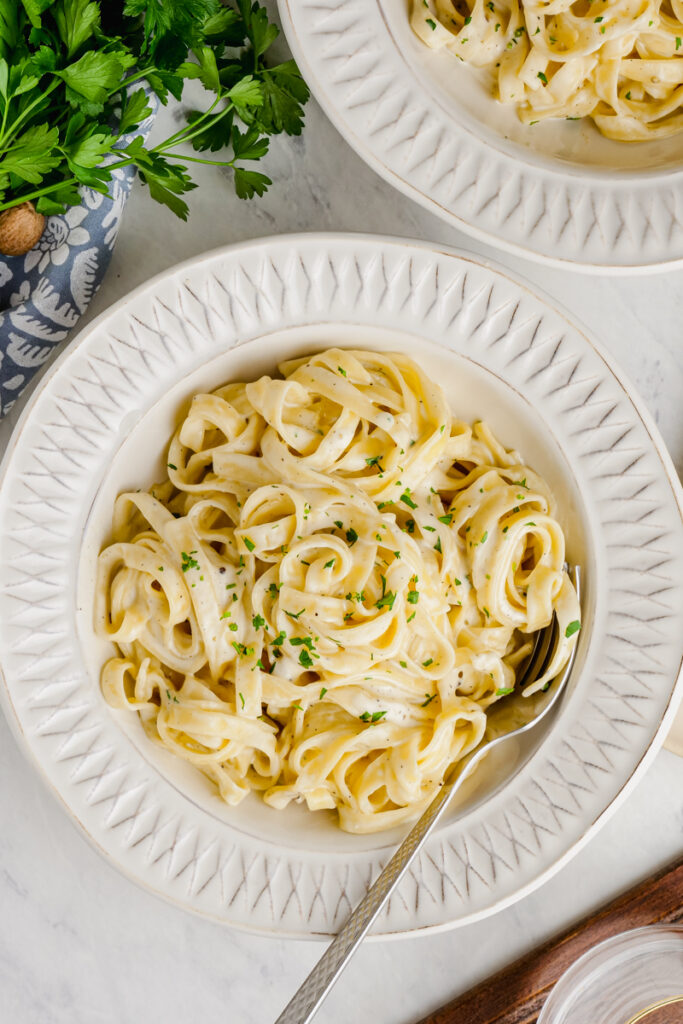 A white plate full of fettuccine alfredo with a fork, and some herbs