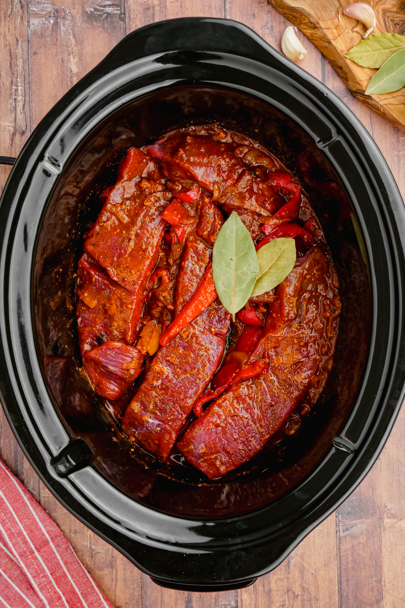 A black slow cooker with all the ingredients for ropa vieja inside, bay leaves on top.