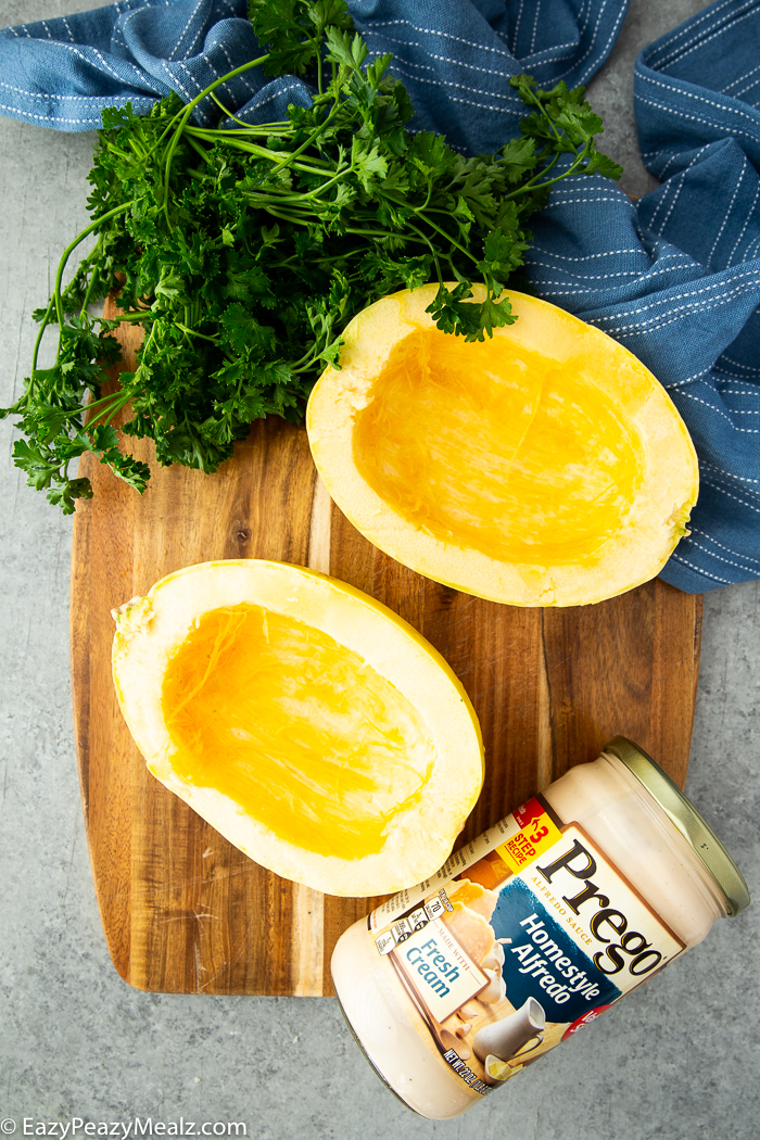 A spaghetti squash cut in half with seeds scraped out, flanked by a jar of Prego, parsley garnish, a blue napkin.