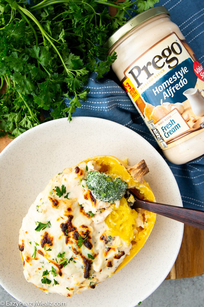 A white bowl with a spaghetti squash stuffed with broccoli chicken alfredo, a wooden fork, and a jar of Prego Homestyle Alfredo