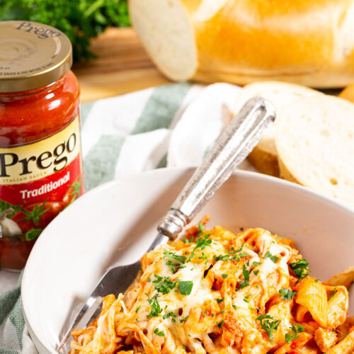 A white bowl with slow cooker chicken parmesan casserole in it, a bottle of Prego Sauce, and some french bread in the background.