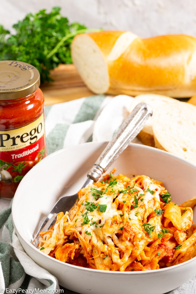 A white bowl with slow cooker chicken parmesan casserole in it, a bottle of Prego Sauce, and some french bread in the background. 