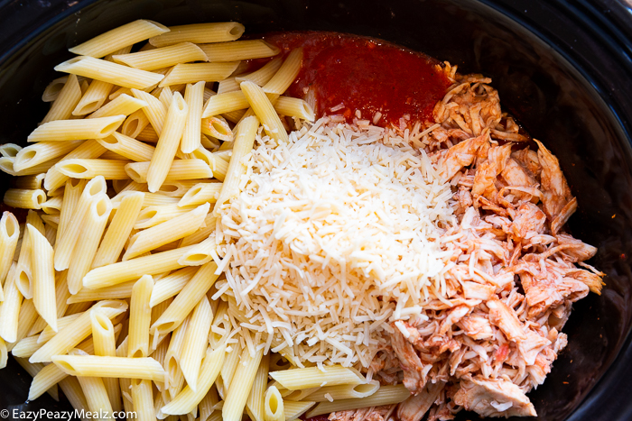 A crock pot with shredded chicken, cheese, and penne.