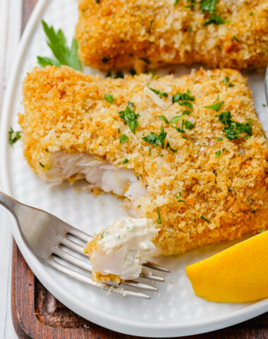 Oven fried fish on a white plate with flakes of it on a fork and lemon wedges