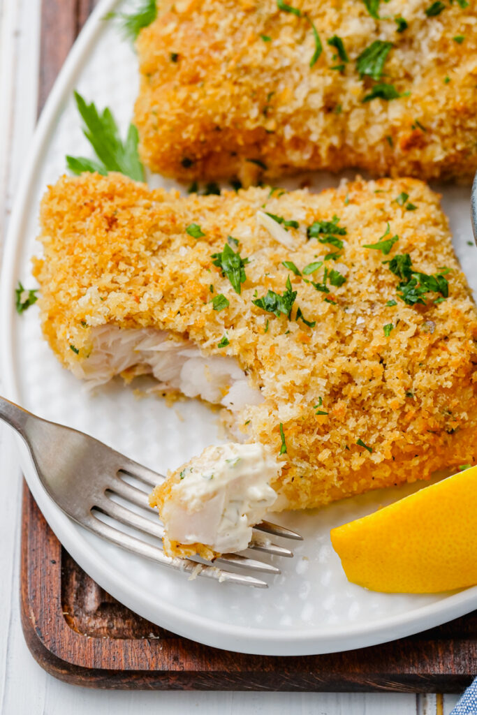 Oven fried fish on a white plate  with flakes of it on a fork and lemon wedges