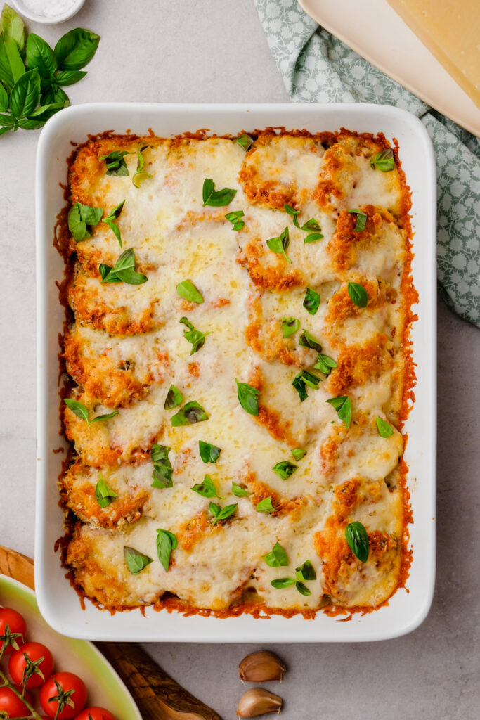 eggplant parmesan dish in white glass container on counter with basil and tomatoes to the side