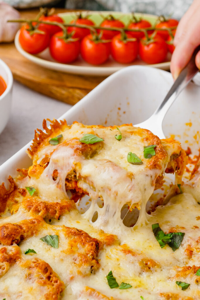 Eggplant parmesan in a white baking dish, being scooped out by a metal spatula, tomatoes in background