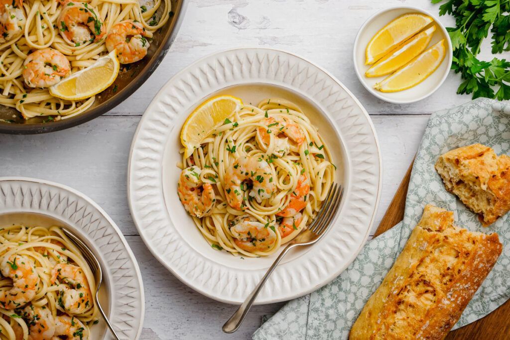 A white plate full of shrimp scampi, with lemon wedges and crusty bread on the side. 