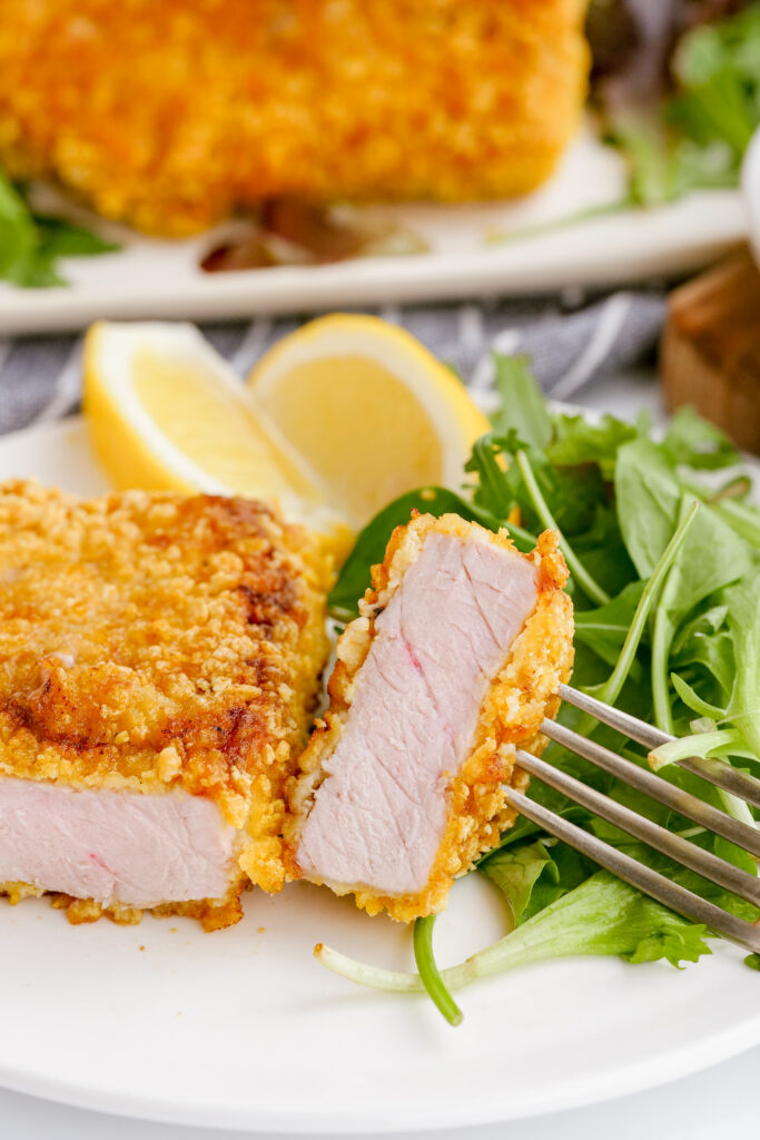 A breaded pork chop on a plate, cut into with a fork and knife, lemon wedges in the background. 