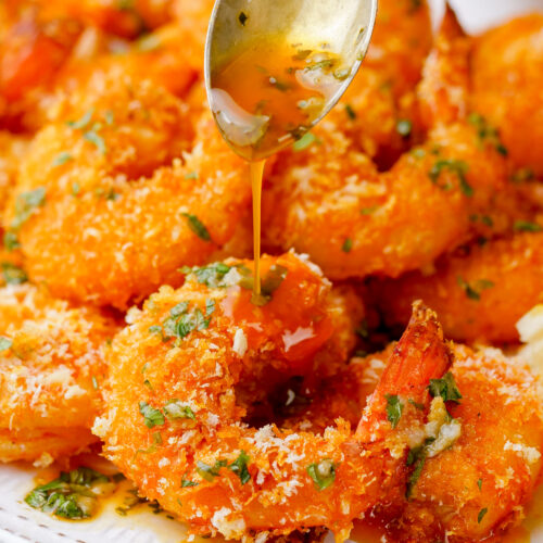 breaded shrimp that is oven baked on a white plate with sauce being spooned over the top.