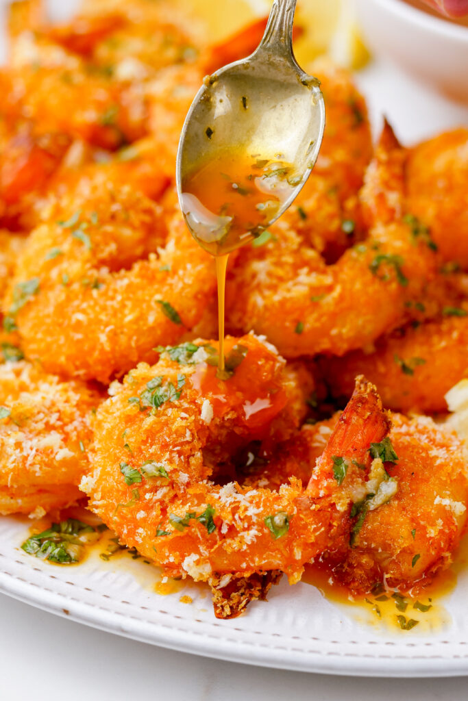 breaded shrimp that is oven baked on a white plate with sauce being spooned over the top. 