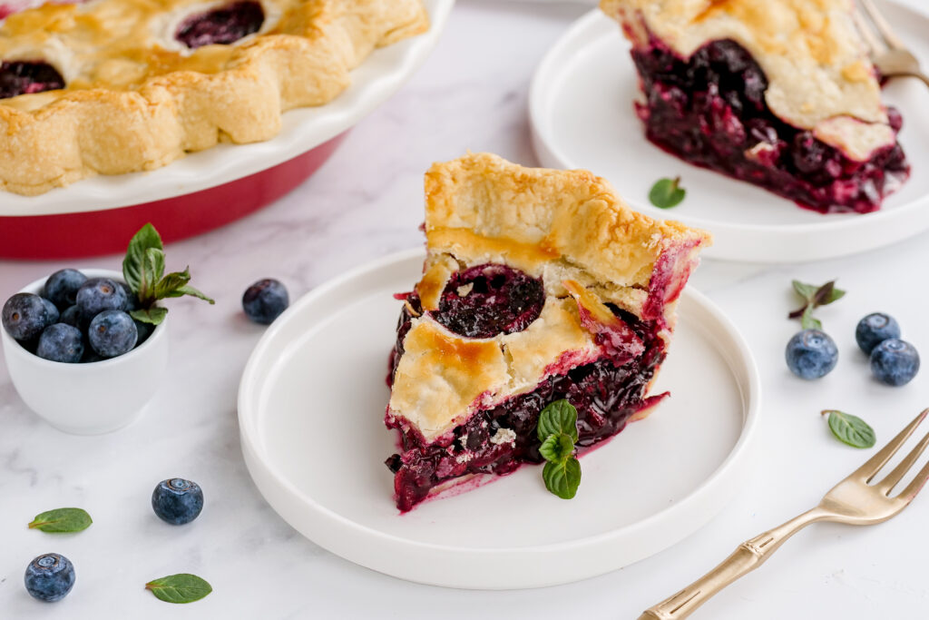 A slice of blueberry pie on a white plate, whole pie in the background