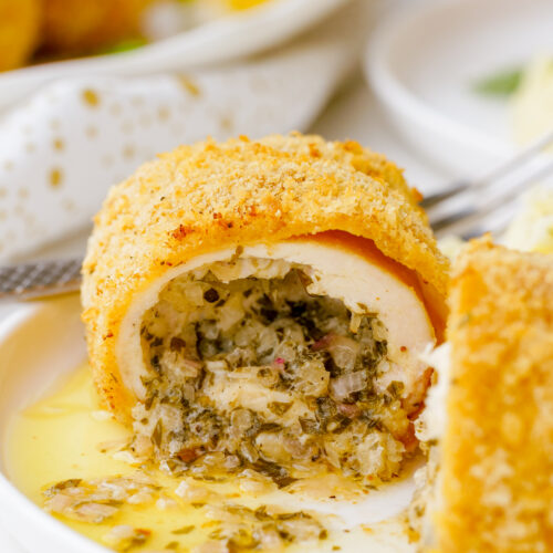 A white plate with a serving of chicken kiev, a golden, delicious, herb butter stuffed chicken
