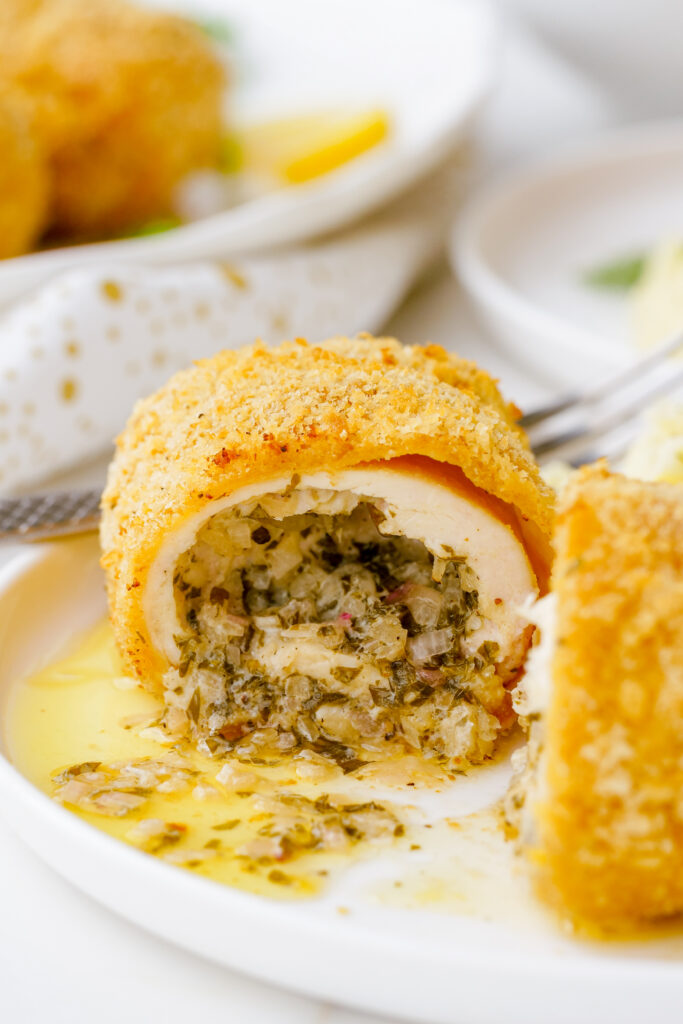 A white plate with a serving of chicken kiev, a golden, delicious, herb butter stuffed chicken