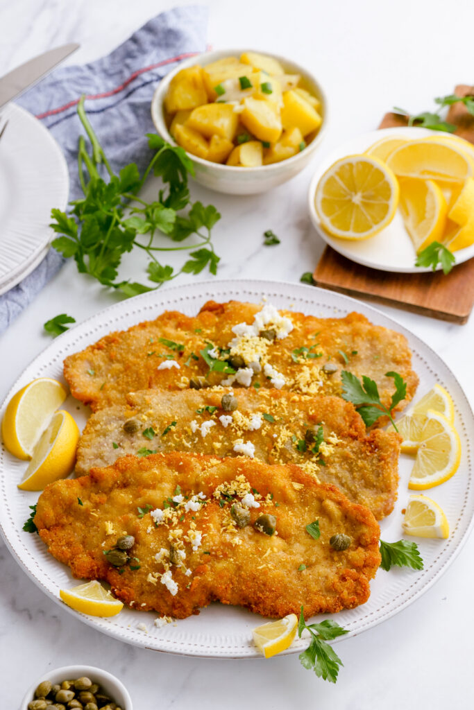 A plate of crispy pork schnitzel with garnishes like lemon, parsley, capers, and hard cooked eggs. 