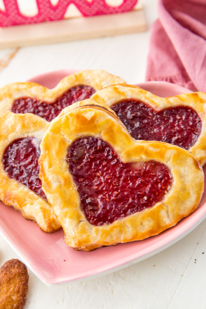 Homemade pop tarts in a heart shape for Valentine's Day, on a cute heart plate. The most fun, 3 ingredients, "Pop Hearts", or in other words, buttery, flakey, raspberry homemade pop tarts. Indulge in these fresh, homemade, but oh so simple, raspberry filled pop tarts! Crispy and flaky on the outside, warm and gooey and tart raspberry on the inside, each bite is bursting with flavor, and they take only minutes to make